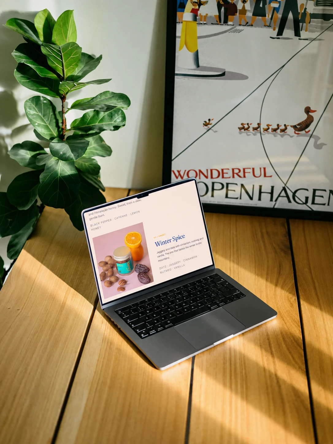 A laptop on a sunlit oak table beside a fiddle-leaf plant and a framed Copenhagen poster, showing the Winter Spice flavour on the store